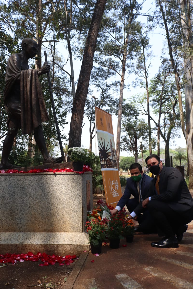 O Secretaria Luiz Alvaro Salles Aguiar de Menezes está agachado ao lado do Consul, em frente à estátua de Gandhi, oferecendo flores. 